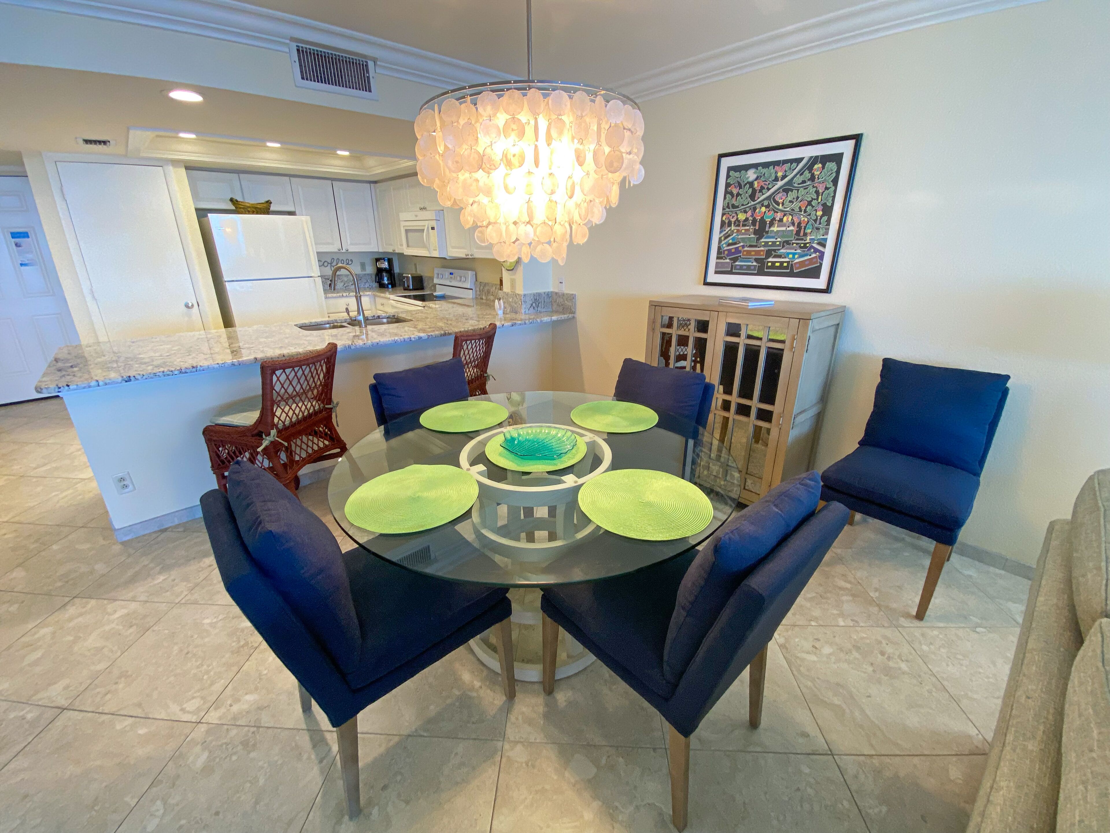 A modern kitchen with white cabinets, a central island, and a dining area with a glass table and blue chairs.