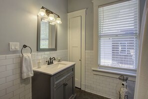 Un baño con tocador blanco, un espejo con tres luces, toallero, dispensador de papel higiénico y una ventana con persianas.