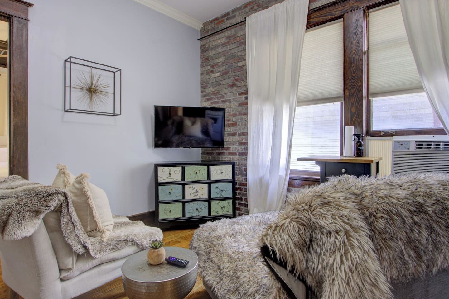 Een woonkamer met een bakstenen muur, een flatscreentelevisie, een decor kast, een zachte bank en een kleine ronde tafel met een afstandsbediening.