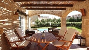 An outdoor patio with a stone wall, a round wooden table, and four cushioned chairs. - Rock House's Villa Melody (2 Bedrooms, Sleeps 4) with private garden and pool. (Platanias)