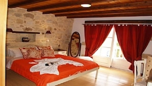 A bedroom with a stone wall, wooden ceiling beams, a bed with red bedding, a bedside table, a mirror, and a window with red curtains. - Rock House's Villa Melody (2 Bedrooms, Sleeps 4) with private garden and pool. (Platanias)