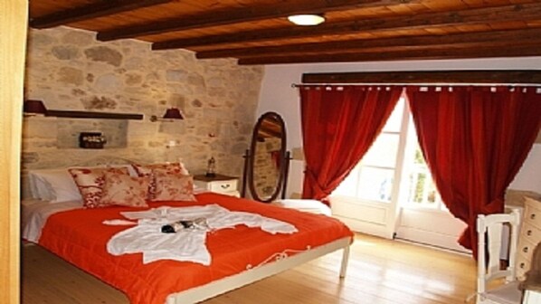 A bedroom with a stone wall, wooden ceiling beams, a bed with red bedding, a bedside table, a mirror, and a window with red curtains. - Rock House's Villa Melody (2 Bedrooms, Sleeps 4) with private garden and pool. (Platanias)