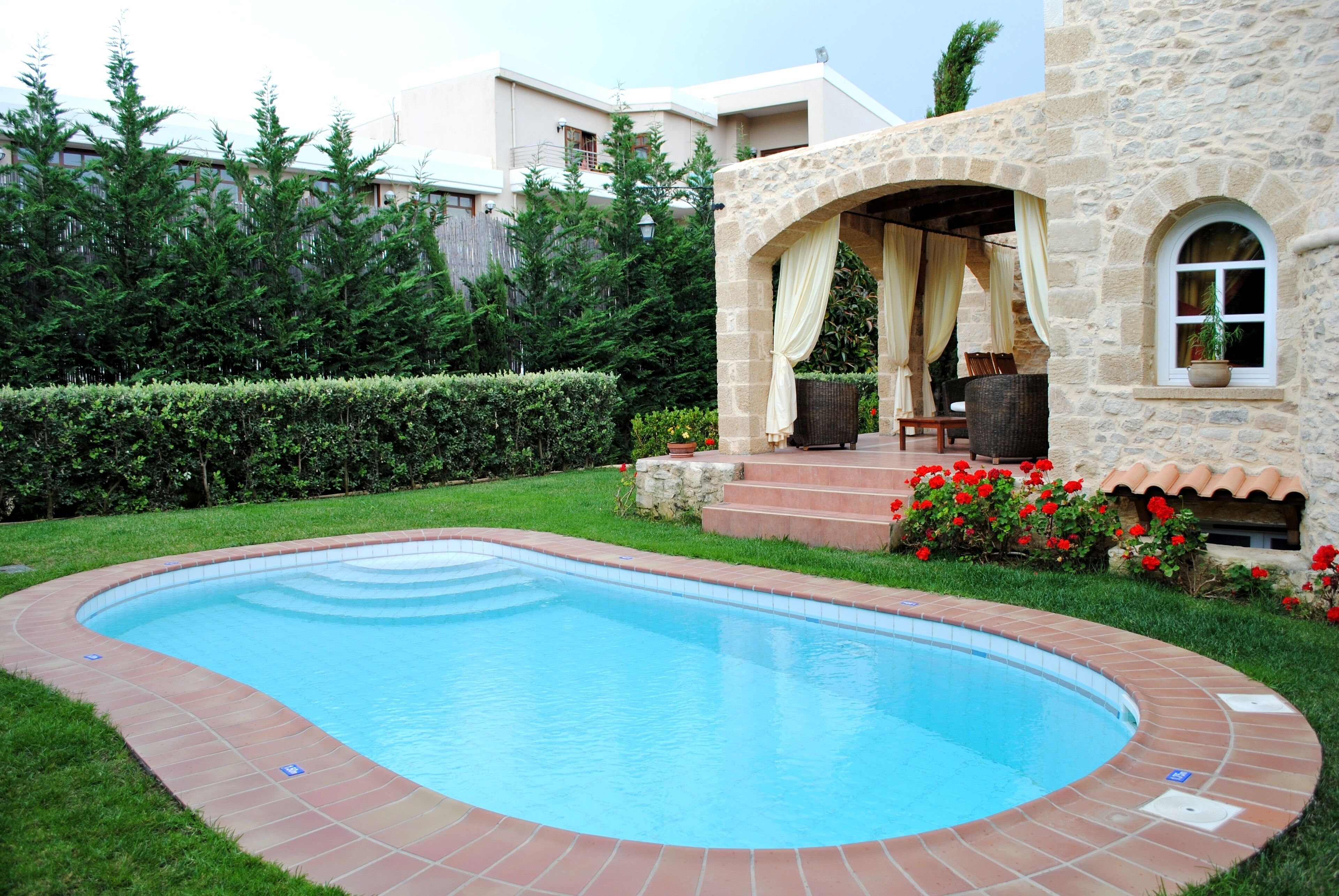 Une piscine avec une terrasse en pierre, un espace salon couvert et un bâtiment en pierre doté d’une fenêtre.