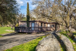 A cabin with a deck, surrounded by trees and a large rock. - Rock View Cabin Property (Oakhurst)