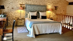 Un dormitorio con pared de piedra, cama con ropa blanca, dos lámparas de noche y una silla de madera.