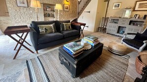 Un salón acogedor con un sofá oscuro, una mesa de centro con libros y una vela, una pared de piedra y una escalera.