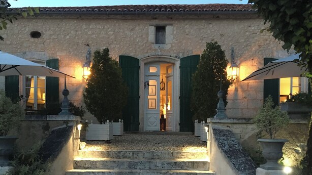 Un edificio de piedra con una puerta central, flanqueado por dos árboles e iluminado con lámparas de pared.