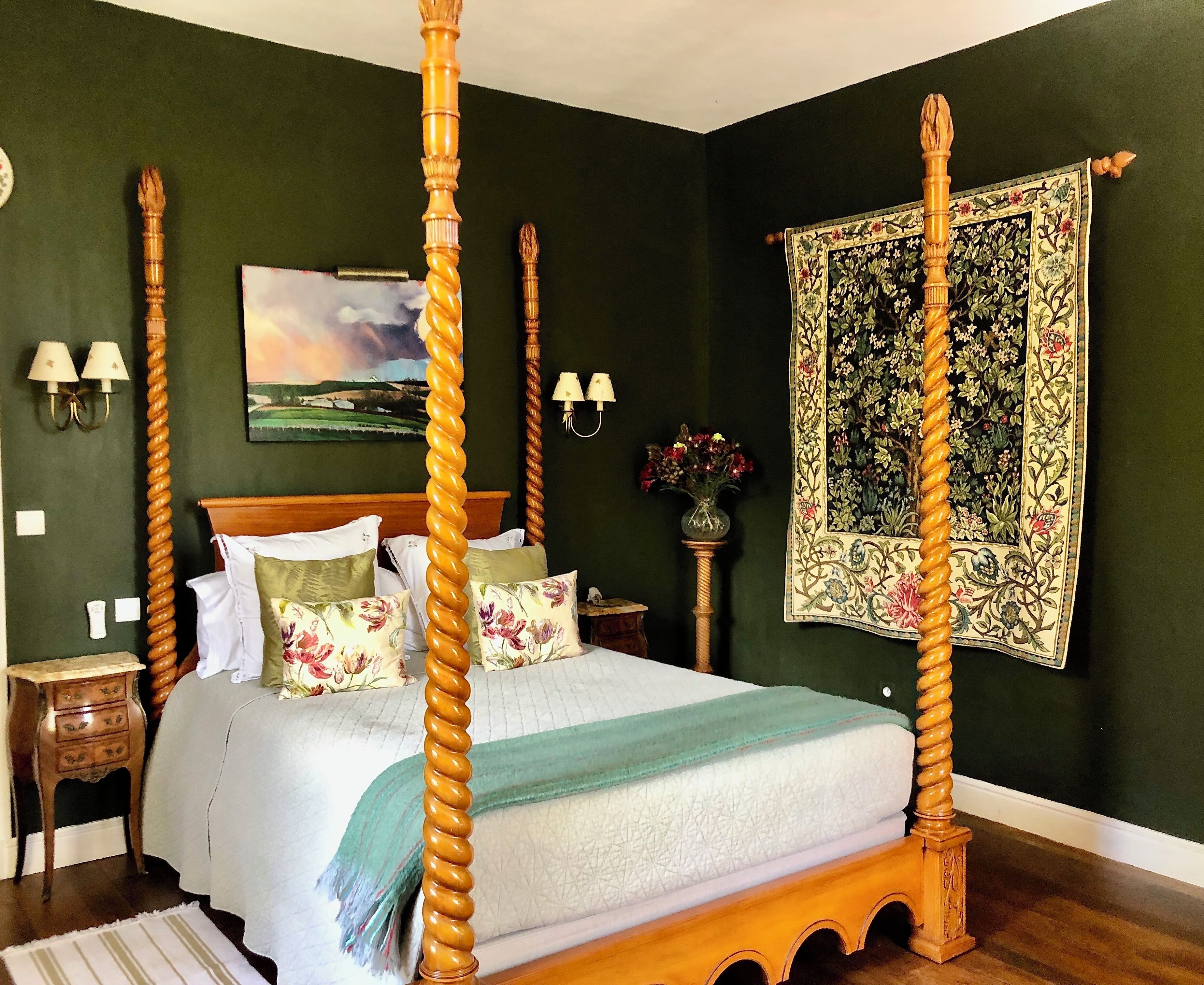 Un lit à baldaquin avec une structure en bois, une couvre-lit à motifs floraux et des coussins assortis. On y trouve une table de chevet avec une lampe, un vase rempli de fleurs et une tapisserie accrochée au mur.