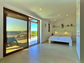 A bedroom with a large sliding glass door leading to a balcony, a bed, a small table, and a wall-mounted shelf with decorative items.