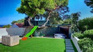 A backyard with artificial grass, a stone planter, a green slide, a swing set, and a car parked near a stone wall. - Charming  Villa, quiet location, stunning view and infinity pool is all you need (Ražanj)