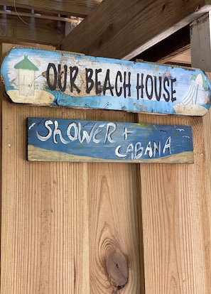 Une pancarte en bois sur laquelle on peut lire « NOTRE MAISON DE PLAGE » et « DOUCHE + CABANE ».