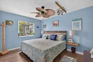A bedroom with a bed, ceiling fan, wooden floor, and artwork on the walls.