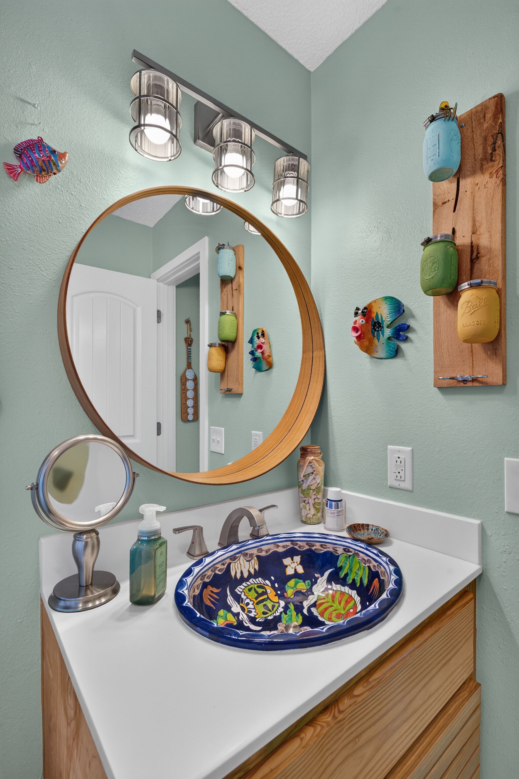 Un baño con un espejo redondo, un lavamanos con un plato decorativo y una repisa de madera con frascos.