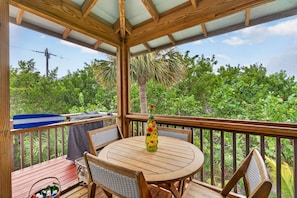 A covered outdoor deck with a round wooden table and chairs, surrounded by lush greenery and a clear sky.
