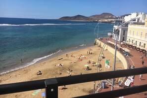 Una playa con gente tomando el sol y nadando, edificios al fondo y un cielo despejado.