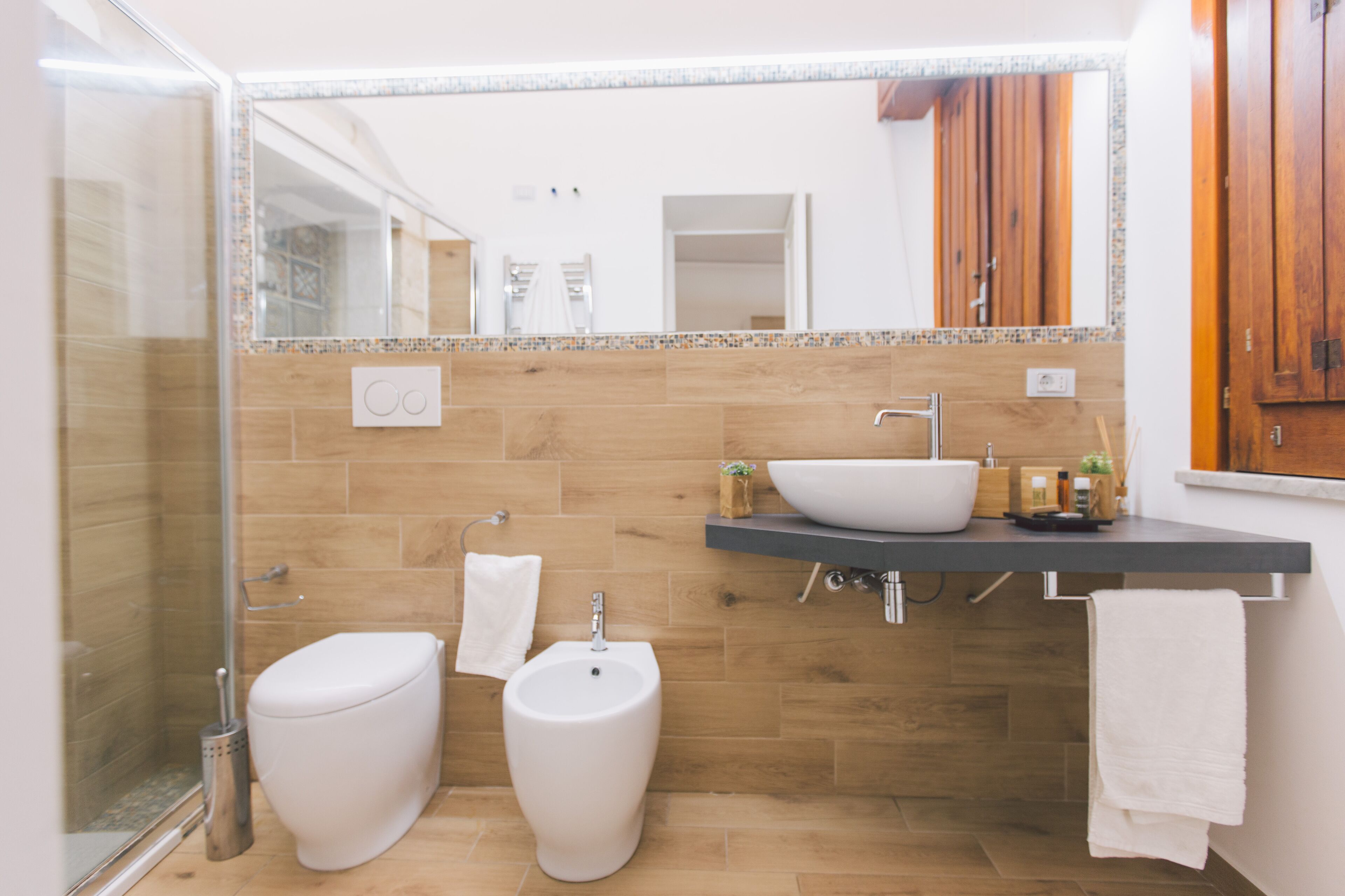 A modern bathroom with a glass shower enclosure, a bidet, a sink with a mirror above, and wooden wall tiles.