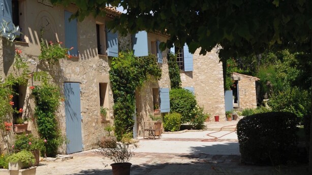 A narrow stone pathway flanked by buildings with blue shutters, surrounded by lush greenery and potted plants.