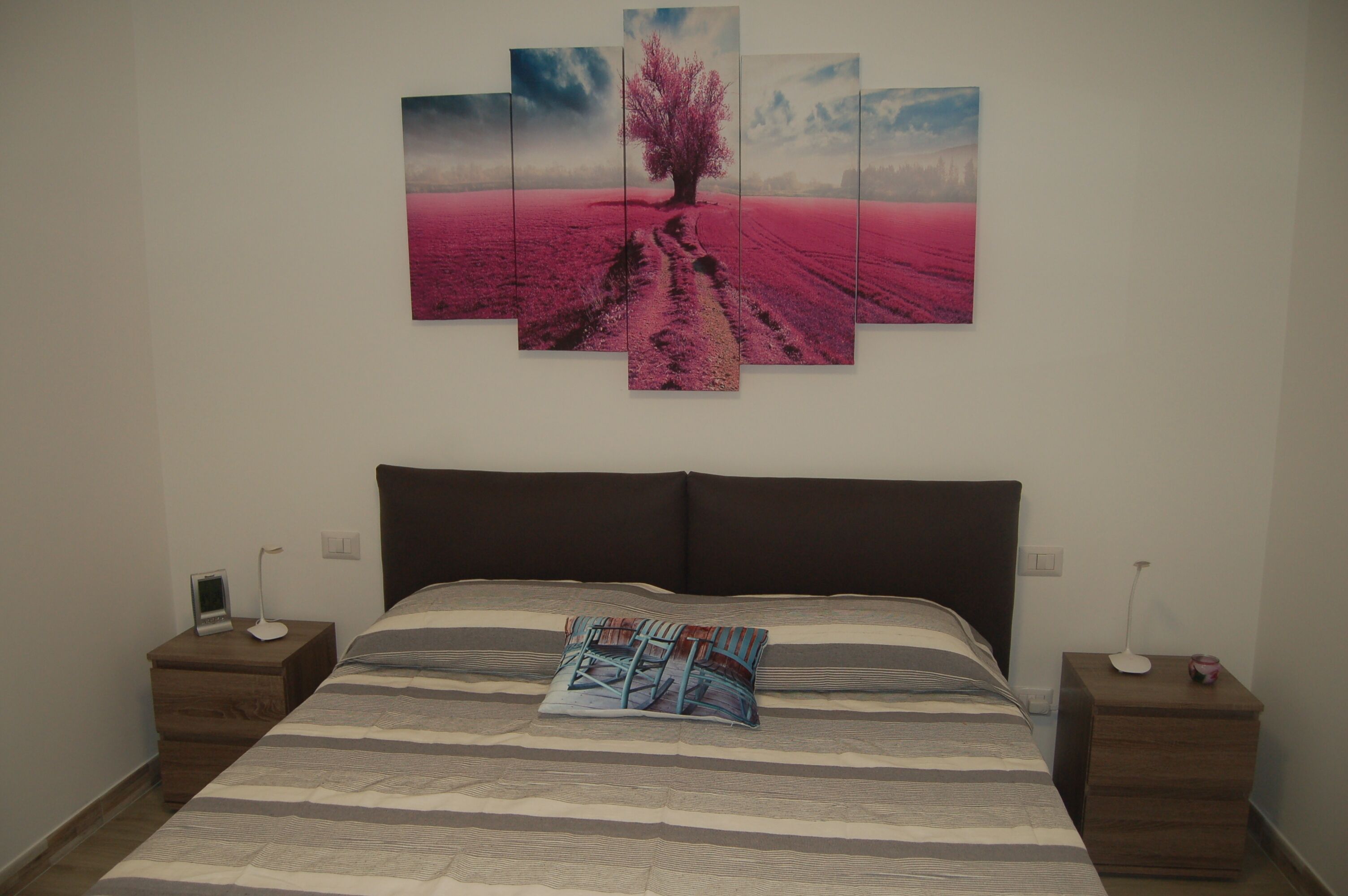 A bedroom with a large bed, a nightstand, a bedside table, and a wall-mounted artwork of a tree on a field.