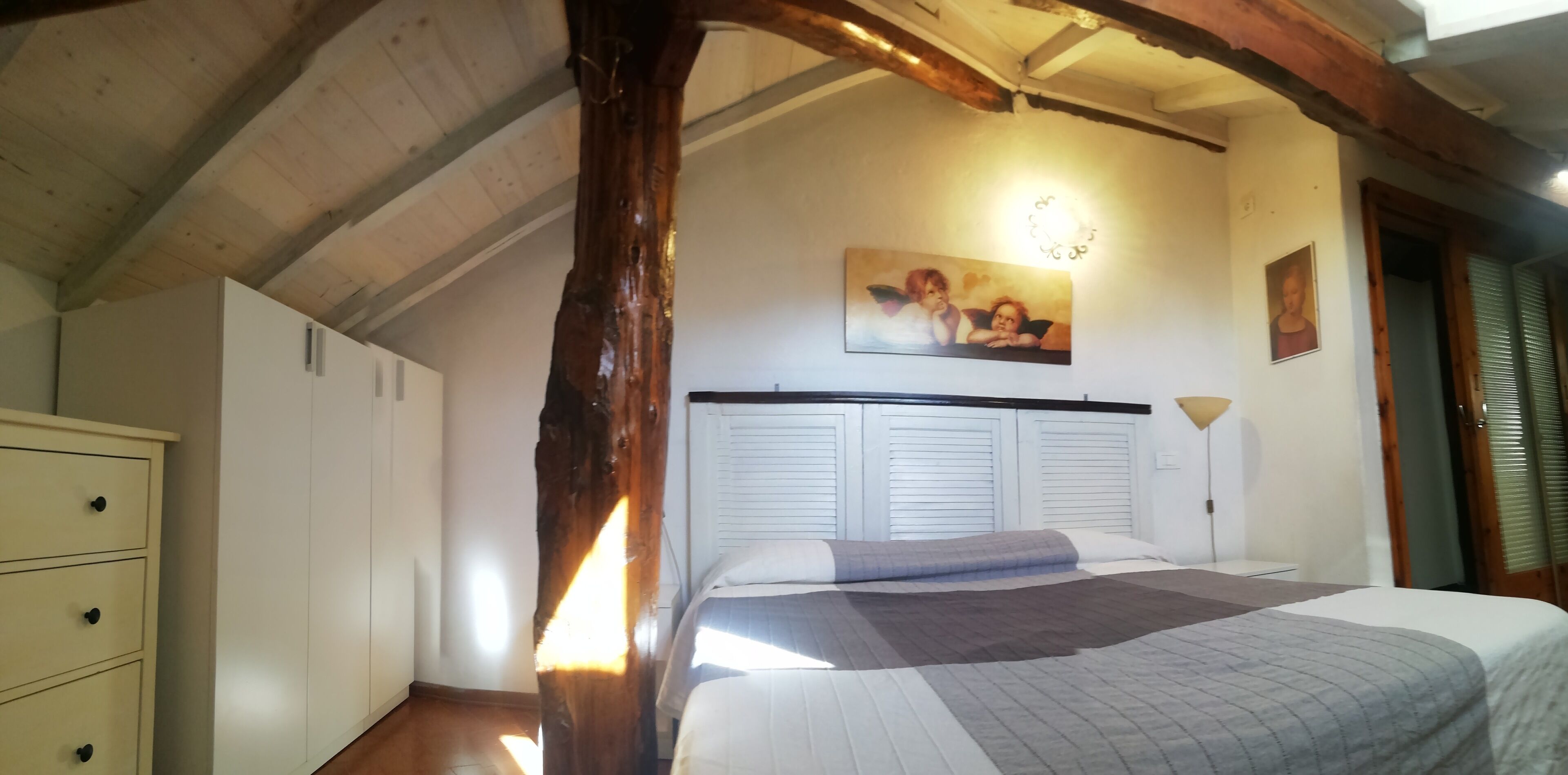A bedroom with a wooden ceiling, a bed with a grey and white duvet, a white wardrobe, and a painting on the wall.