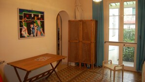 Um quarto com uma mesa de madeira, um armário de vime, um quadro e uma janela com cortinas.