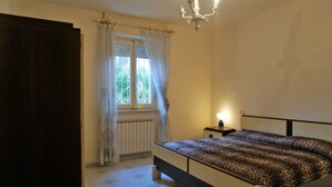 Quarto com cama, mesa de cabeceira, janela com cortinas, radiador e lustre.