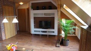 Un salon moderne avec un téléviseur, un plancher en bois, une table en verre ornée de fleurs et une plante en pot.