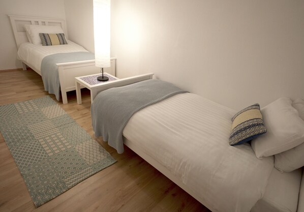 A bedroom with two single beds, a bedside table with a lamp, a patterned rug, and a wooden floor. - Beach House 4 - creative space for creative people (Porches)