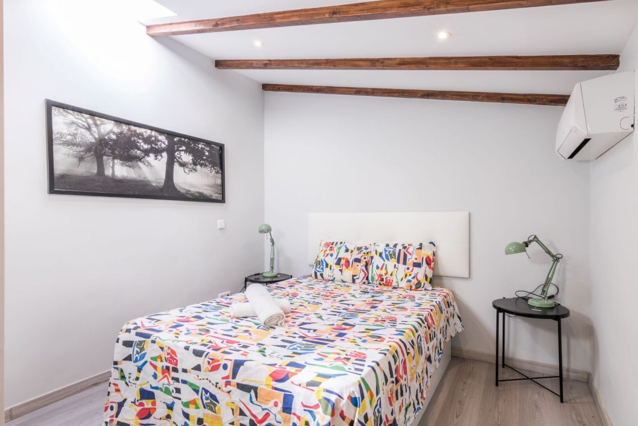 A bedroom with a bed, a nightstand, a desk, a chair, a picture on the wall, and a ceiling fan.