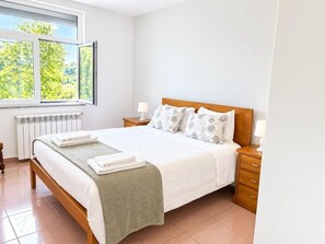 A bedroom with a bed, a nightstand, a lamp, a window with a view of trees, and a radiator.