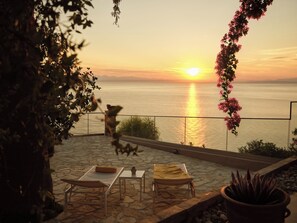 A patio with a view of the sunset over the water, a table with a plant, and a chair.