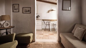 Een gezellige woonkamer met een groene fauteuil, een beige bank en een kleine tafel met een lamp. Een doorgang leidt naar een keuken met een houten plafond en een hangende decoratie.