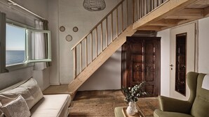 Een kamer met een houten trap, een witte muur, een houten deur, een groene fauteuil, een witte bank, een raam met gordijnen en een hanglamp.