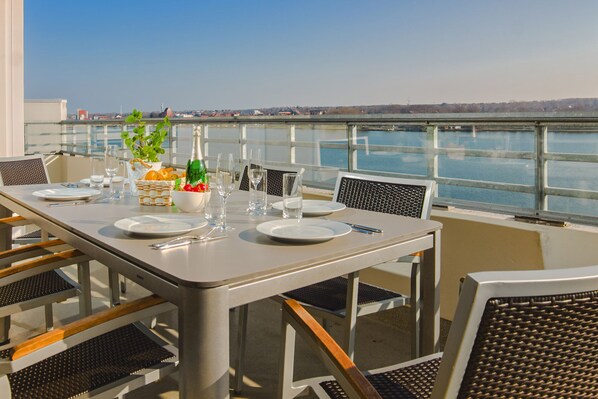 A dining table set with plates, glasses, and a bottle, overlooking a body of water with a bridge in the background. - Design penthouse apartment on the steinwarder with its own sauna (Heiligenhafen)