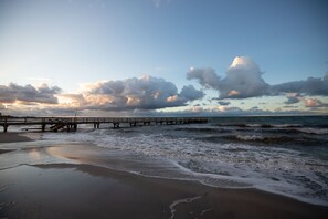 A beach with a wooden pier extending into the ocean, waves gently lapping at the shore, and a partly cloudy sky. - Design penthouse apartment on the steinwarder with its own sauna (Heiligenhafen)