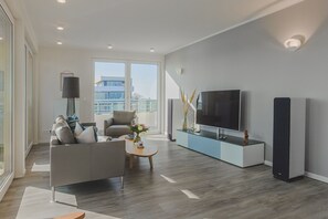 A modern living room with a grey sofa, a coffee table, a TV on a blue stand, and a large window offering a city view. - Design penthouse apartment on the steinwarder with its own sauna (Heiligenhafen)