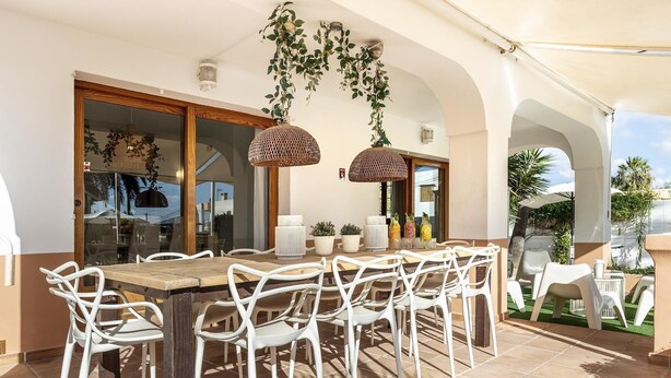 An outdoor dining area with a wooden table and white chairs, surrounded by potted plants and hanging plants. - 🌴 Villa Kiku Ibiza – Pool and pre-party room · 8 min Ushuaïa (Sant Josep de sa Talaia)