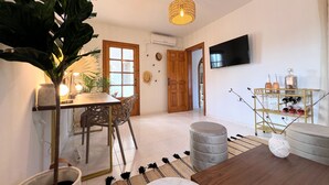 A modern living room with a wooden dining table, chairs, a large potted plant, a hanging light fixture, a TV mounted on the wall, and a glass display case with bottles. - 🌴 Villa Kiku Ibiza – Pool and pre-party room · 8 min Ushuaïa (Sant Josep de sa Talaia)