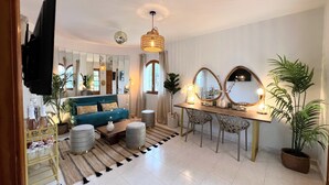 A spacious living room with a blue sofa, a wooden coffee table, and a dining area with a wooden table and chairs. There are two large mirrors on the wall, a chandelier, and several potted plants. - 🌴 Villa Kiku Ibiza – Pool and pre-party room · 8 min Ushuaïa (Sant Josep de sa Talaia)