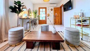 A modern living room with a wooden coffee table, two grey ottomans, a dining area with chairs and a plant, and a TV mounted on the wall. - 🌴 Villa Kiku Ibiza – Pool and pre-party room · 8 min Ushuaïa (Sant Josep de sa Talaia)