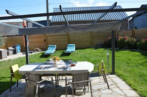 Un patio con mesa y sillas, una zona de estar cubierta y un jardín con flores y una valla.