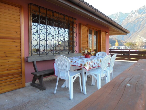 Un edificio de madera con una mesa y sillas afuera, una ventana con rejas decorativas y una vista de la montaña al fondo.