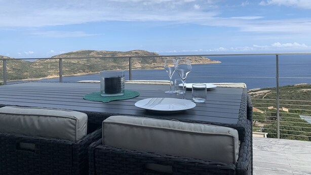 Et bord med et glaslys, to vinglas og en tallerken, beliggende på en balkon med udsigt over et kystlandskab.