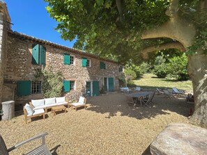 Un bâtiment en pierre doté d’une terrasse en gravier, de sièges extérieurs et d’un arbre qui offre de l’ombre.