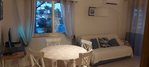 A room with a round table and chairs, a sofa with cushions, a window with curtains, and a wall-mounted lamp.