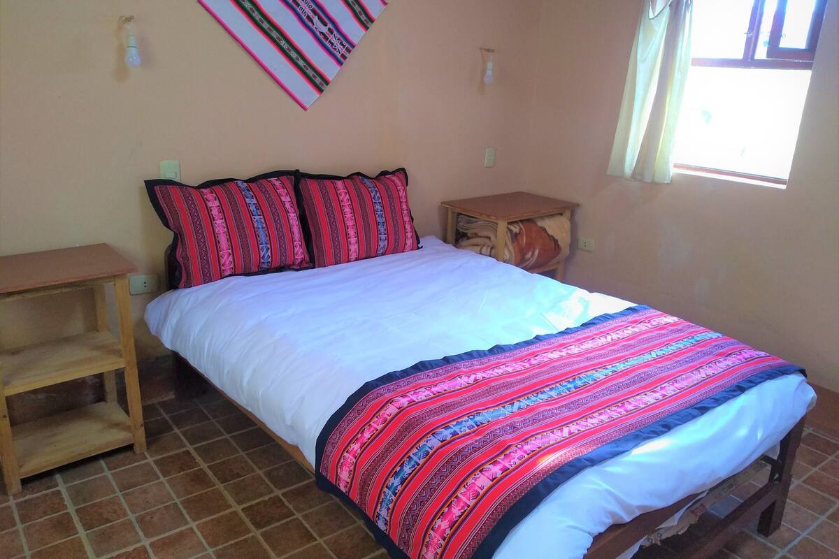 A bed with a white sheet and a colorful blanket, a wooden nightstand, a wall-mounted light, and a window with curtains.