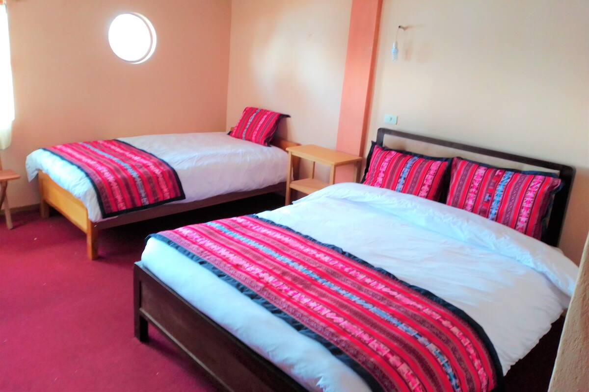 A room with two single beds, each with a patterned bedspread and pillows, a small wooden table, and a round window.