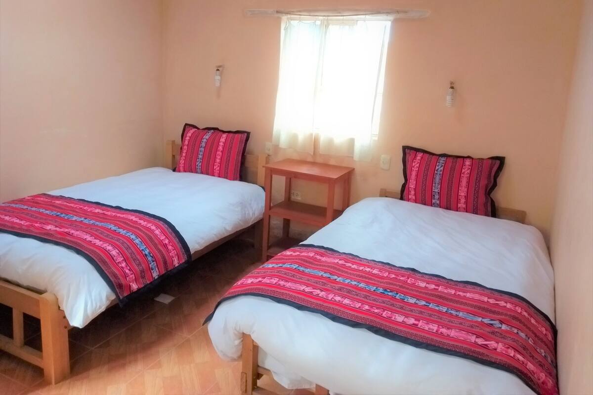 A bedroom with two single beds, each with a patterned blanket and pillows, a small wooden nightstand, and a window with curtains.