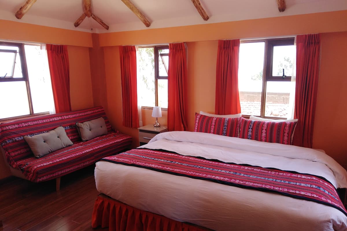 A room with a large bed, a sofa, and two windows with red curtains.