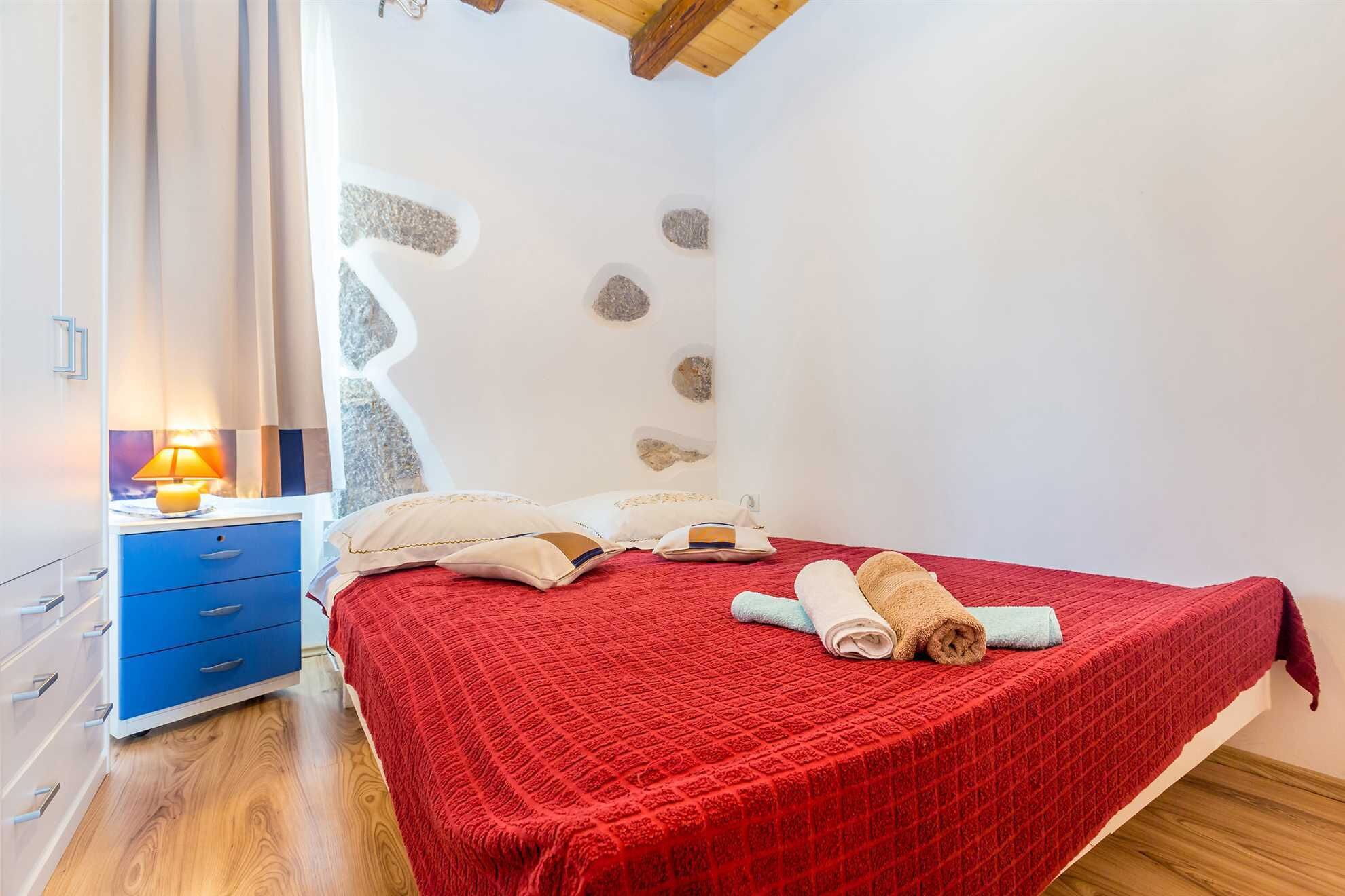 Ein Schlafzimmer mit roter gesteppter Bettdecke, Holzboden, Nachttisch mit Lampe und Wand mit steinartigen Verzierungen.