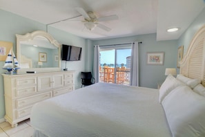 A bedroom with a large bed, a ceiling fan, a dresser, a TV, and a balcony with a view.